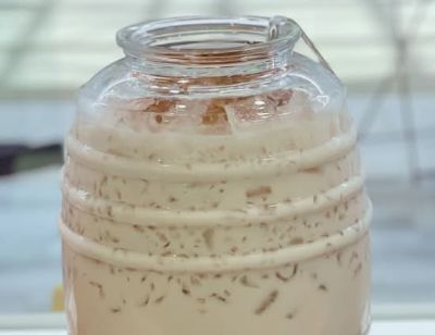 A glass barrel jar filled with a creamy, light brown drink and ice
